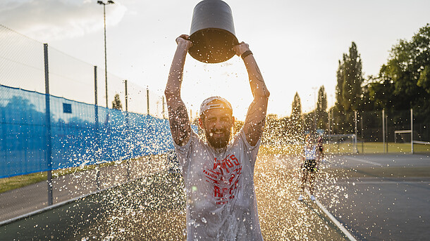 Mann kippt sich auf dem Tennisplatz einen Eimer Wasser über den Kopf.